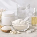 Colagénio hidrolisado em várias formas numa imagem em tons brancos, com colher de pau com pó, frasco com pó branco, óleo em frasco e comprimidos e cápsulas.